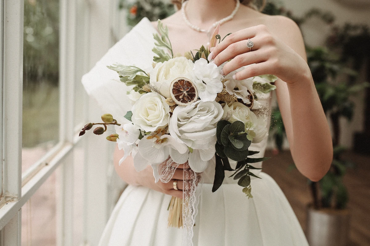 Bride holding an elegant white bouquet with roses, orchids, and eucalyptus, wearing a classic white gown and pearl necklace.