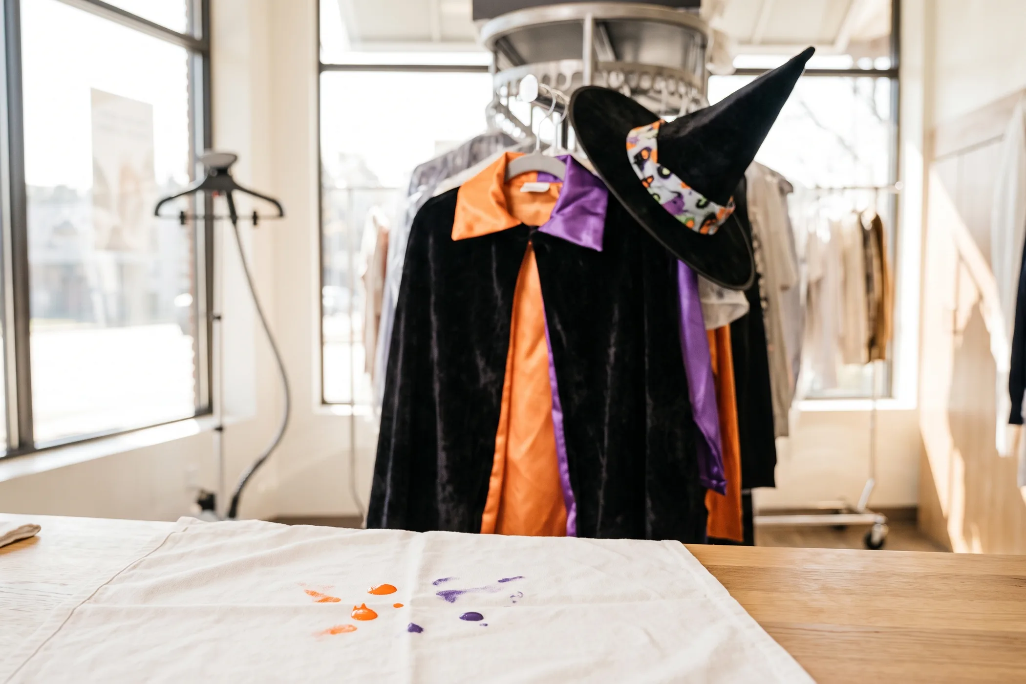 A child's black cape and witch hat on a rack at a dry cleaner with face paint on a white cloth